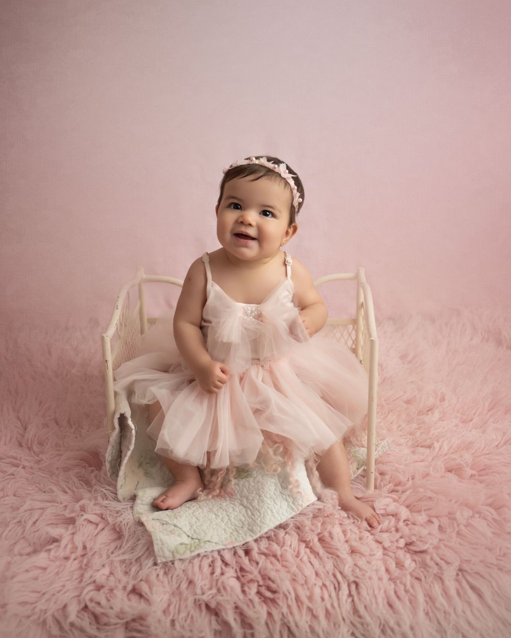 Baby in tulle dress explores sitting position on pink furry rug with white wooden prop.