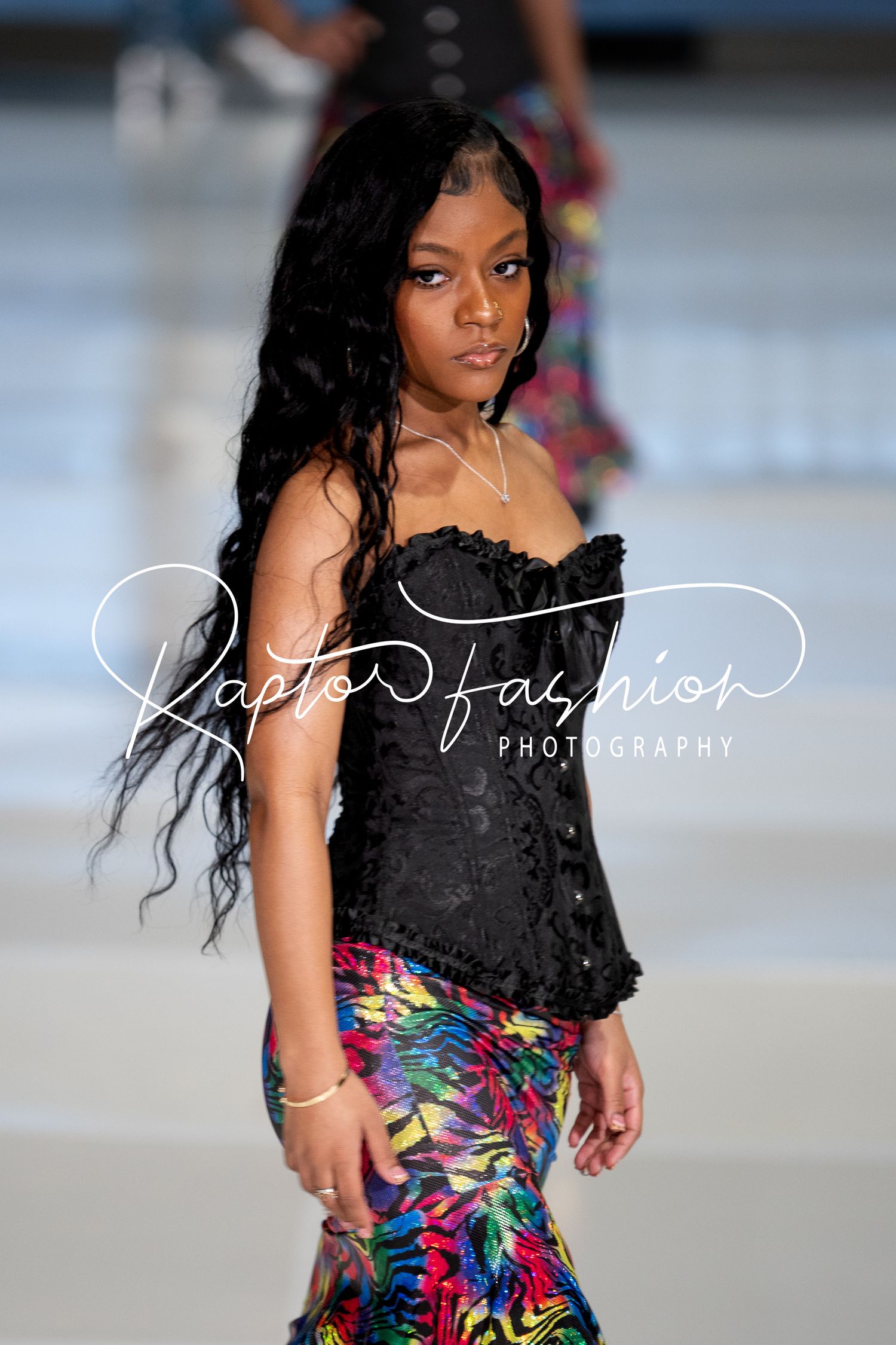 Model walks runway in black corset top with colorful floral print fitted skirt during fashion show.