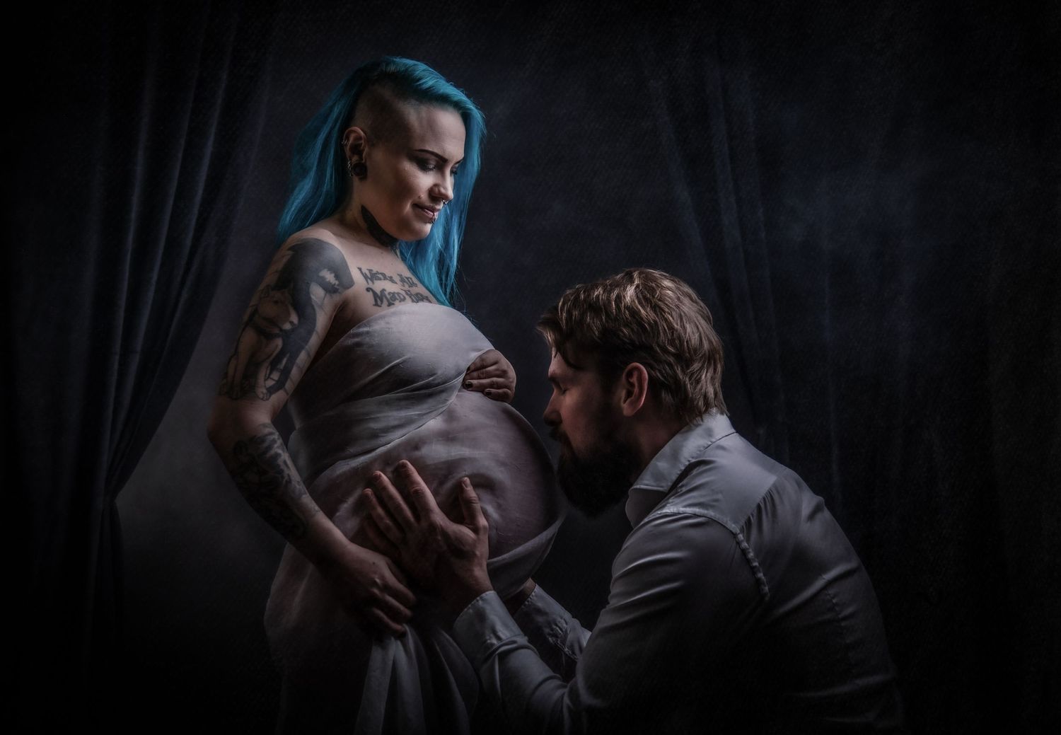 A moody maternity photo with dramatic lighting shows an intimate moment between expectant parents in a dark setting.