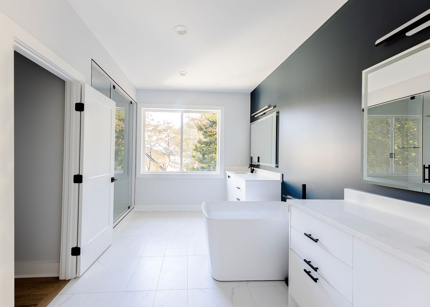 Modern bathroom with white freestanding tub, navy accent wall, white vanity, and large window offering natural light.