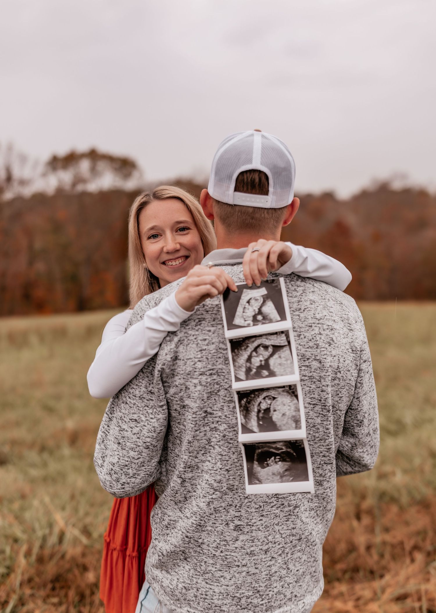 A couple in a field sharing ultrasound photos pinned to the back of a gray sweater during a pregnancy announcement.