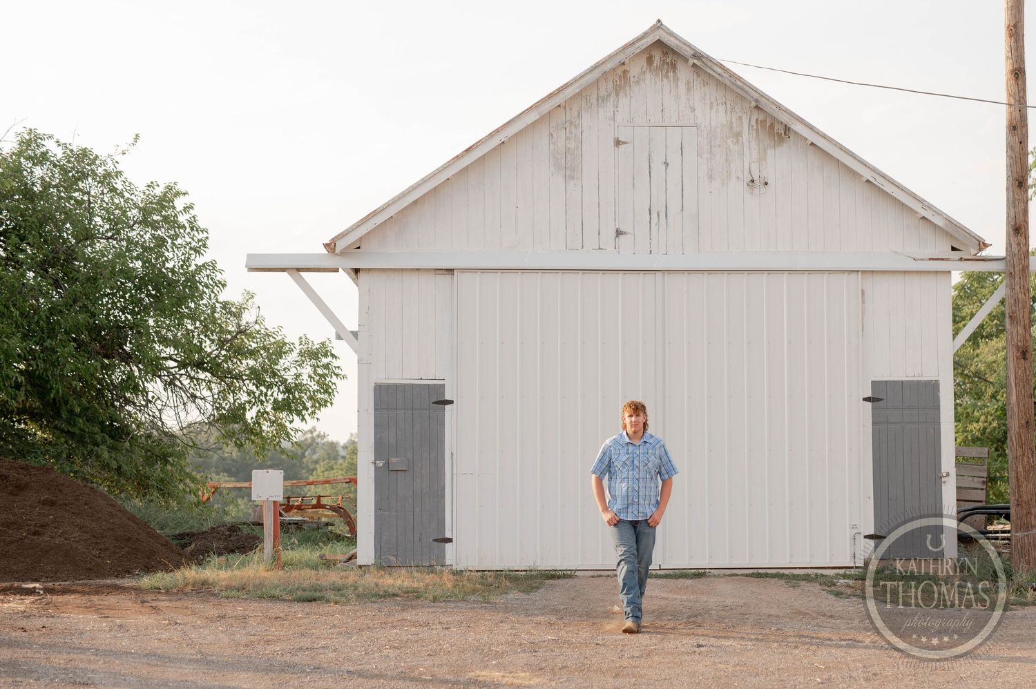 A figure walks past a large white barn with gray doors in a rustic farm setting.