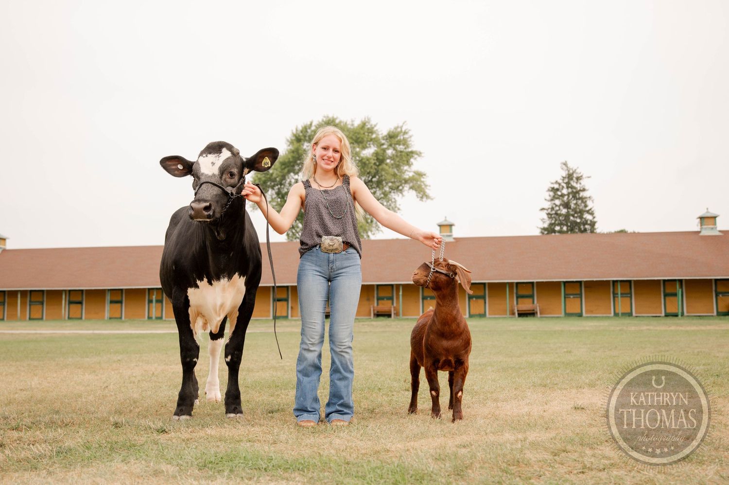 Young person standing between a large black and white cow and a brown calf in front of farm buildings.