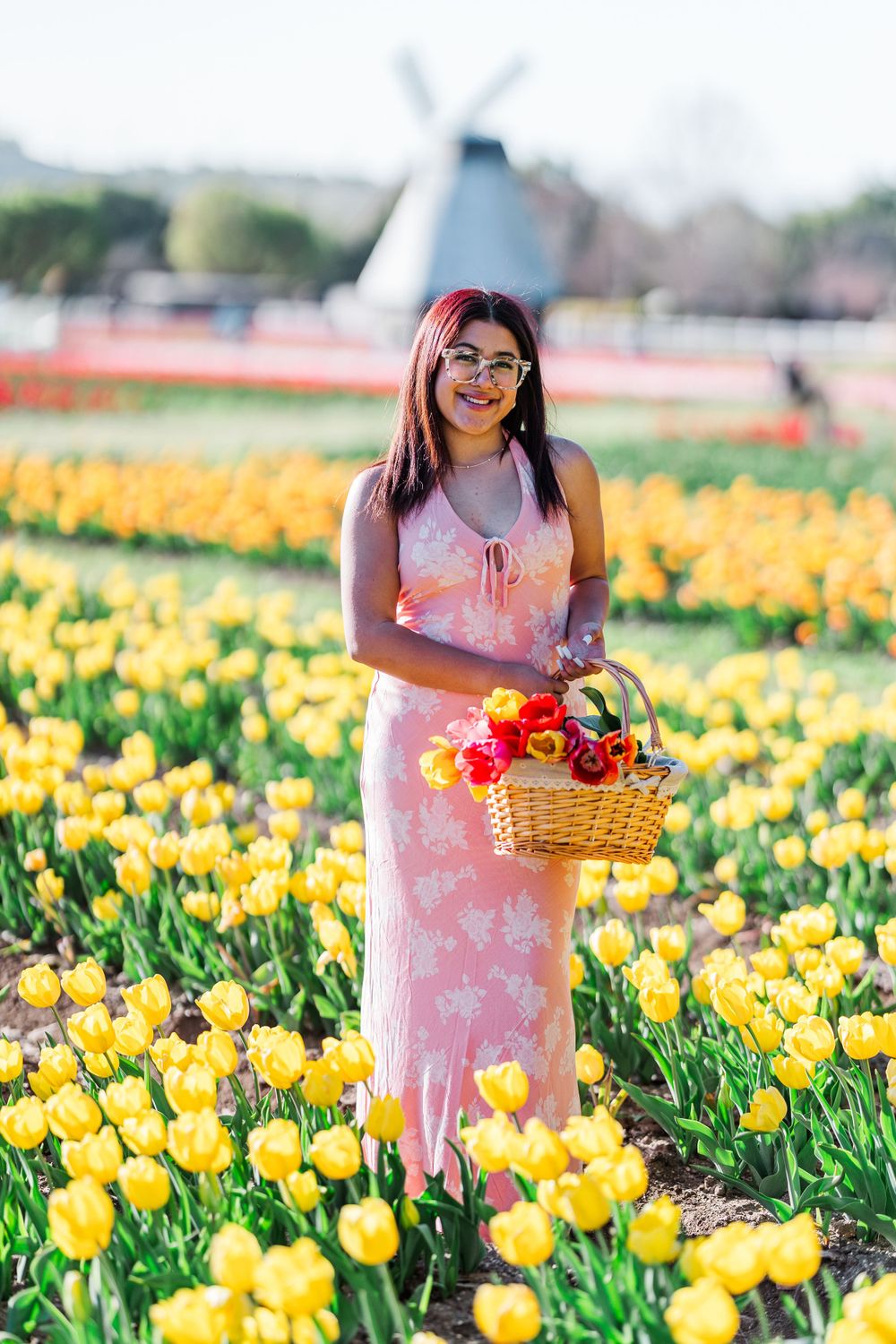 Person in pink floral dress stands among vibrant yellow tulips holding basket of colorful flowers in spring garden field.