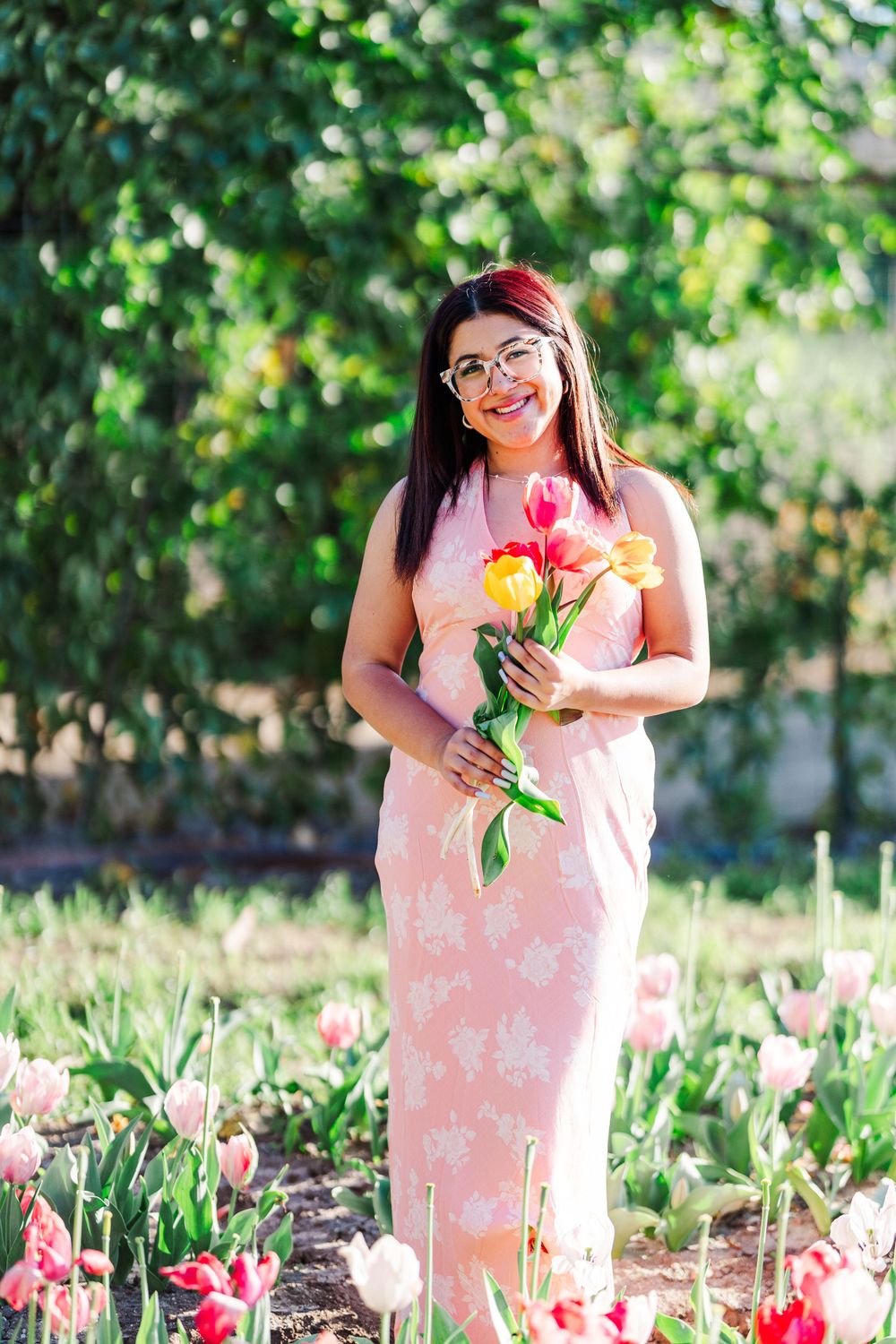 A person in a pink floral dress holds tulips while standing in a sunny garden with blooming flowers.