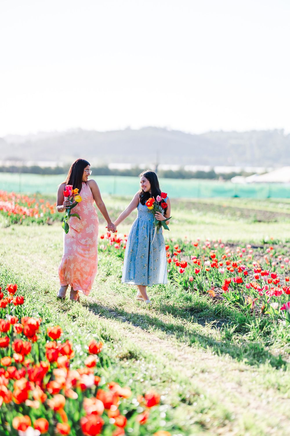 Pair of friends walking through field of red tulips in floral dresses on sunny day.