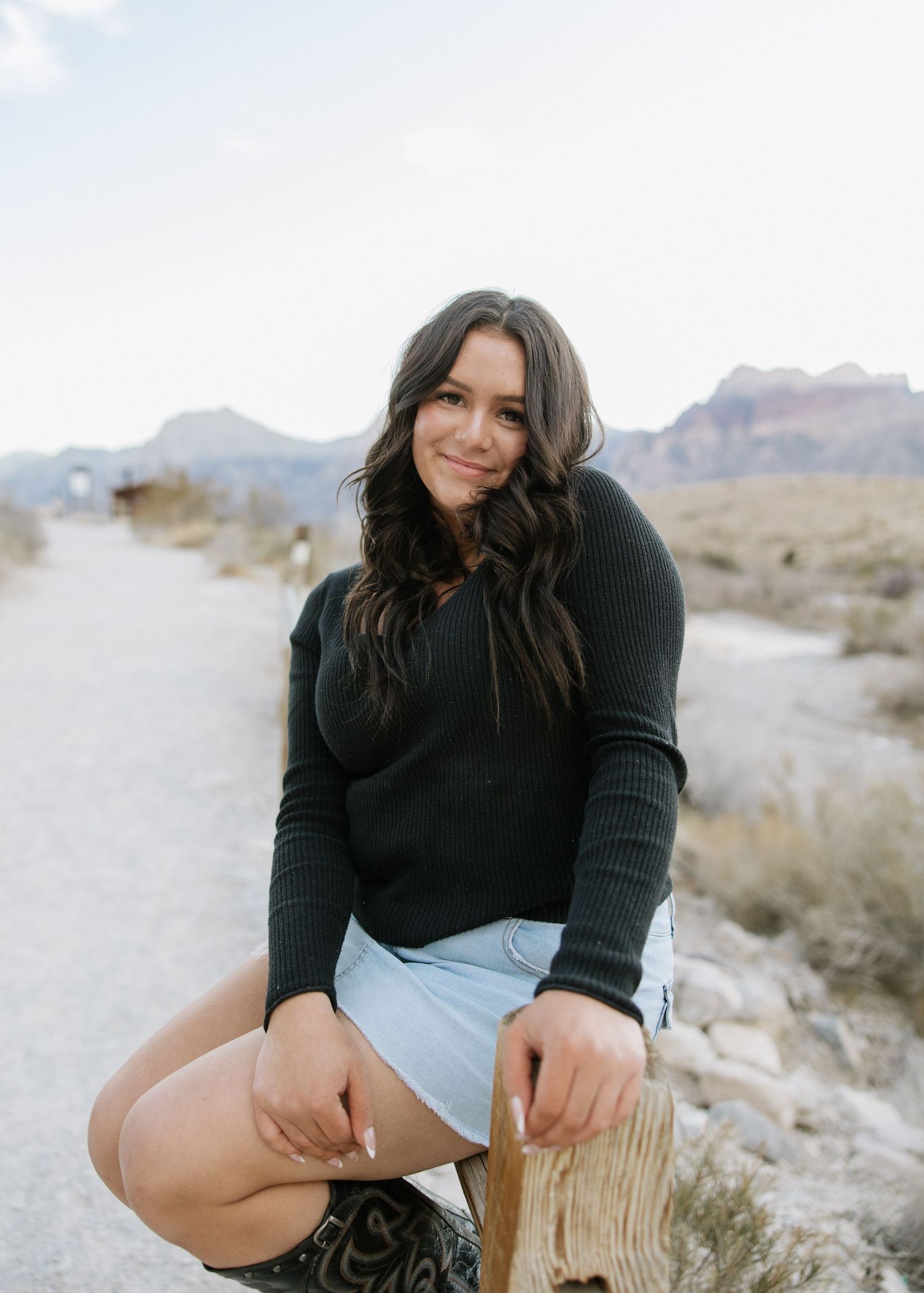 Someone in a black sweater sitting on a wooden log against a desert mountain backdrop on a sunny day.