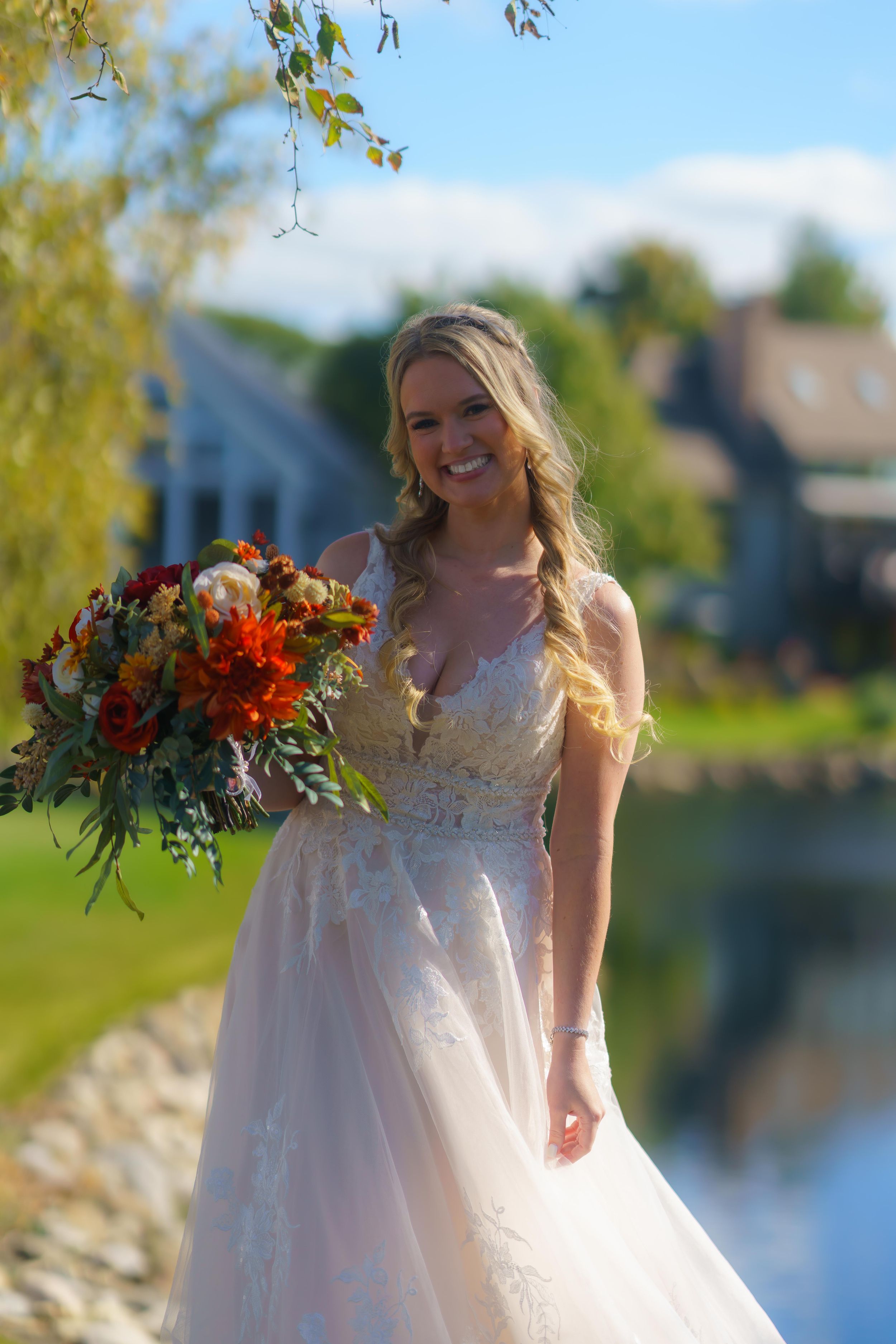 A bride in a flowing white dress poses with vibrant orange and red fall bouquet by a lakeside.