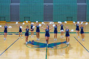 Cheerleaders perform synchronized routine formations on a gymnasium floor with blue and green decor.