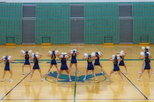 Cheerleaders perform synchronized dance routine in formation on a gymnasium floor with green walls.