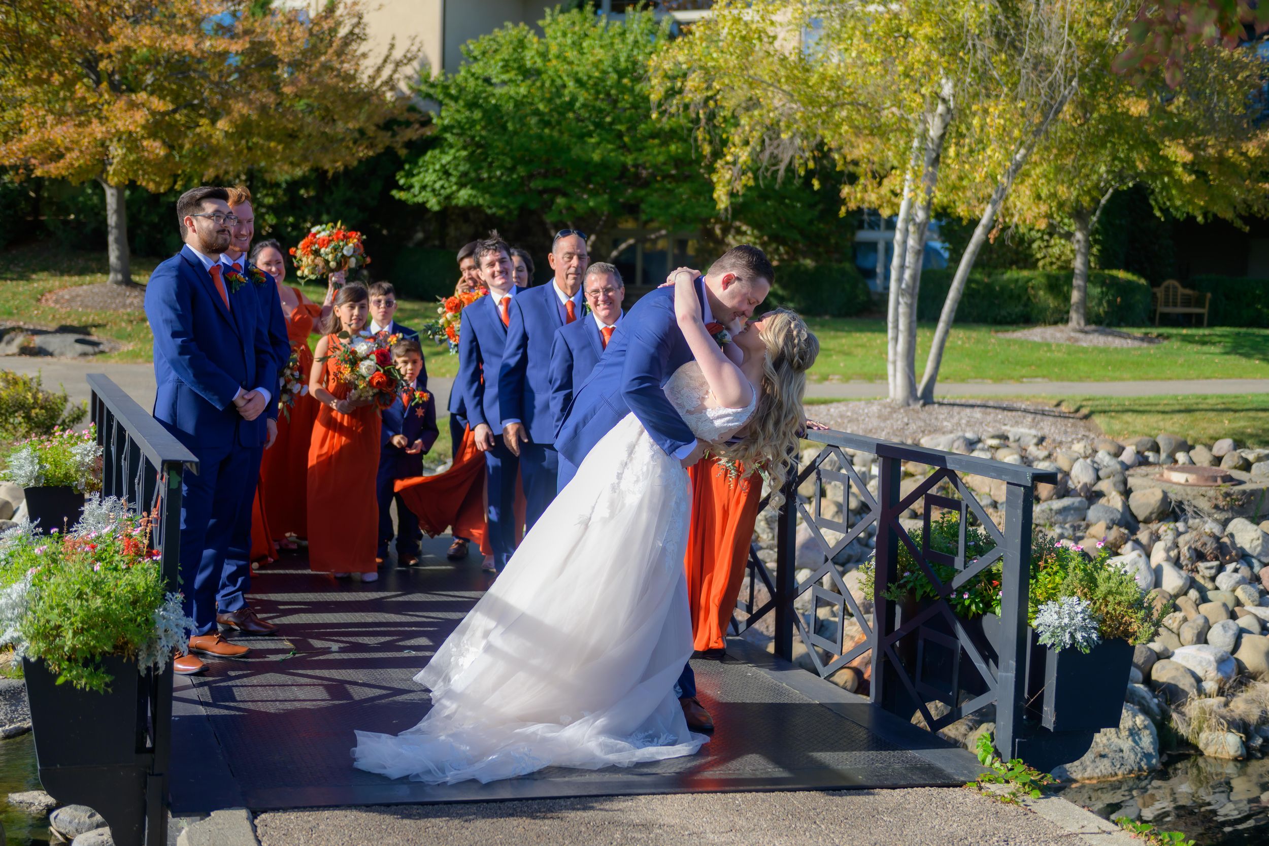 Outdoor wedding ceremony with guests gathered along stone path surrounded by autumn foliage.