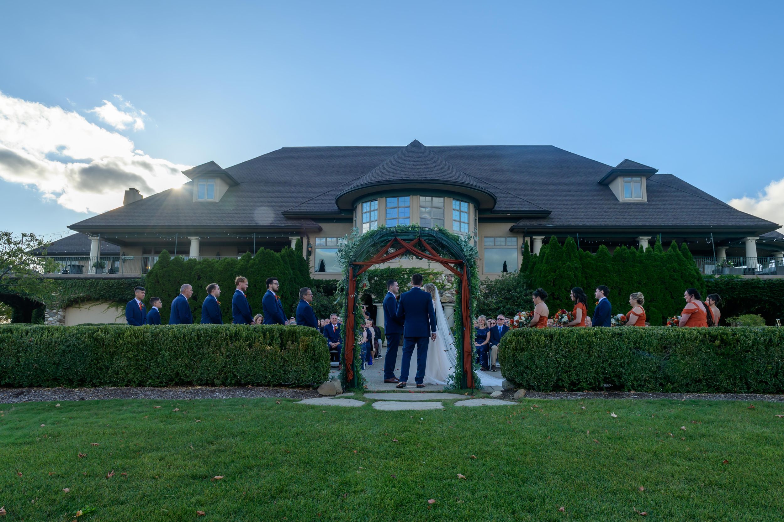 Wedding ceremony taking place under an archway at an elegant estate with manicured hedges and a grand building facade.