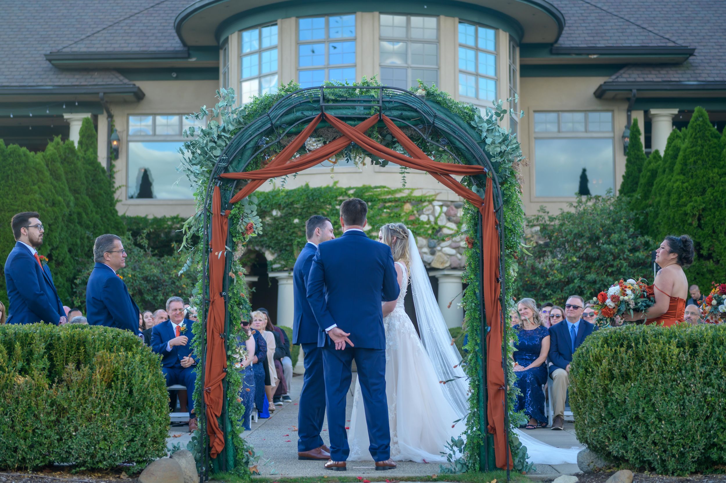 A wedding ceremony takes place under a red gazebo surrounded by lush gardens and greenery.