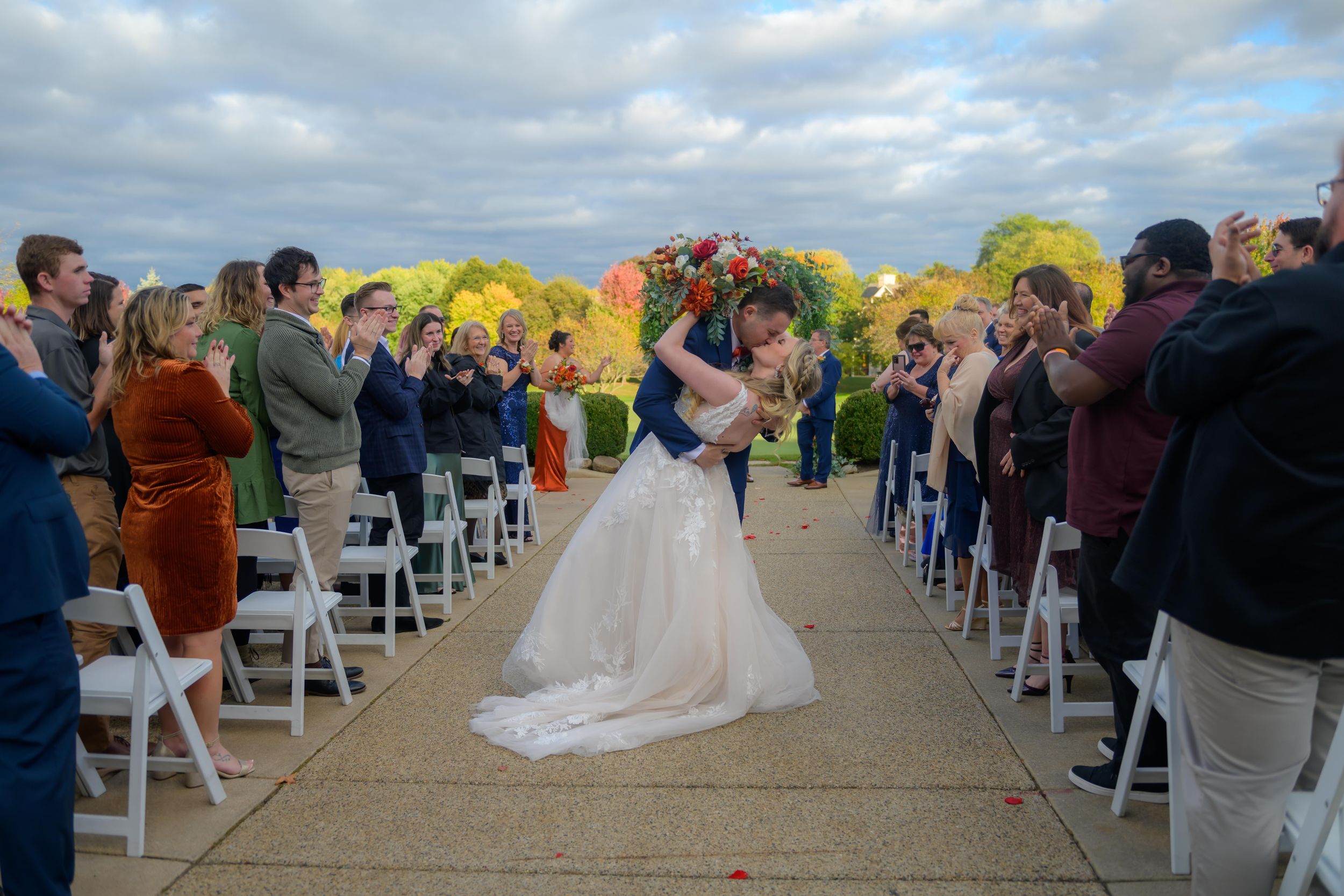 Wedding ceremony panorama with guests seated in white chairs against scenic fall landscape.