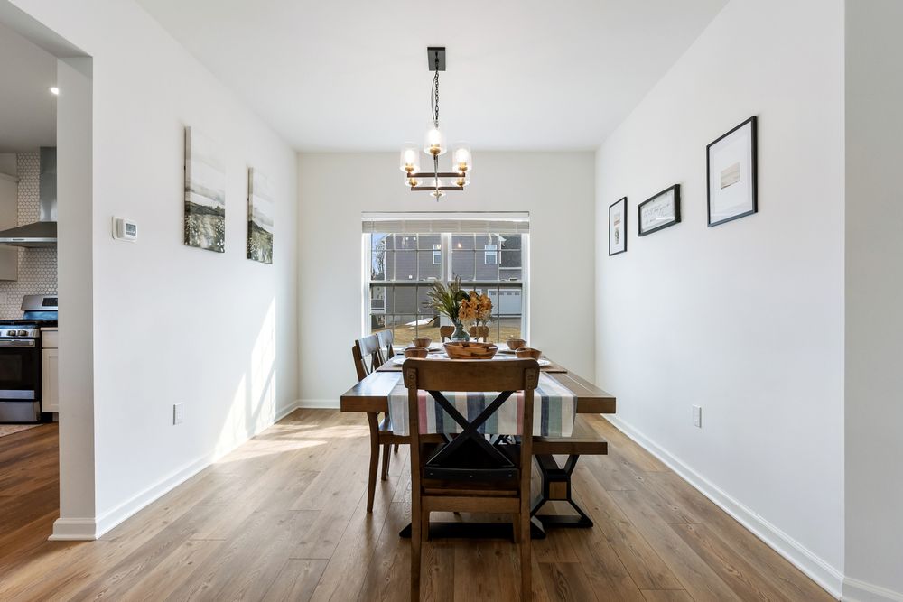 Bright dining room with hardwood floors, rustic wooden table, and modern chandelier near a large window.