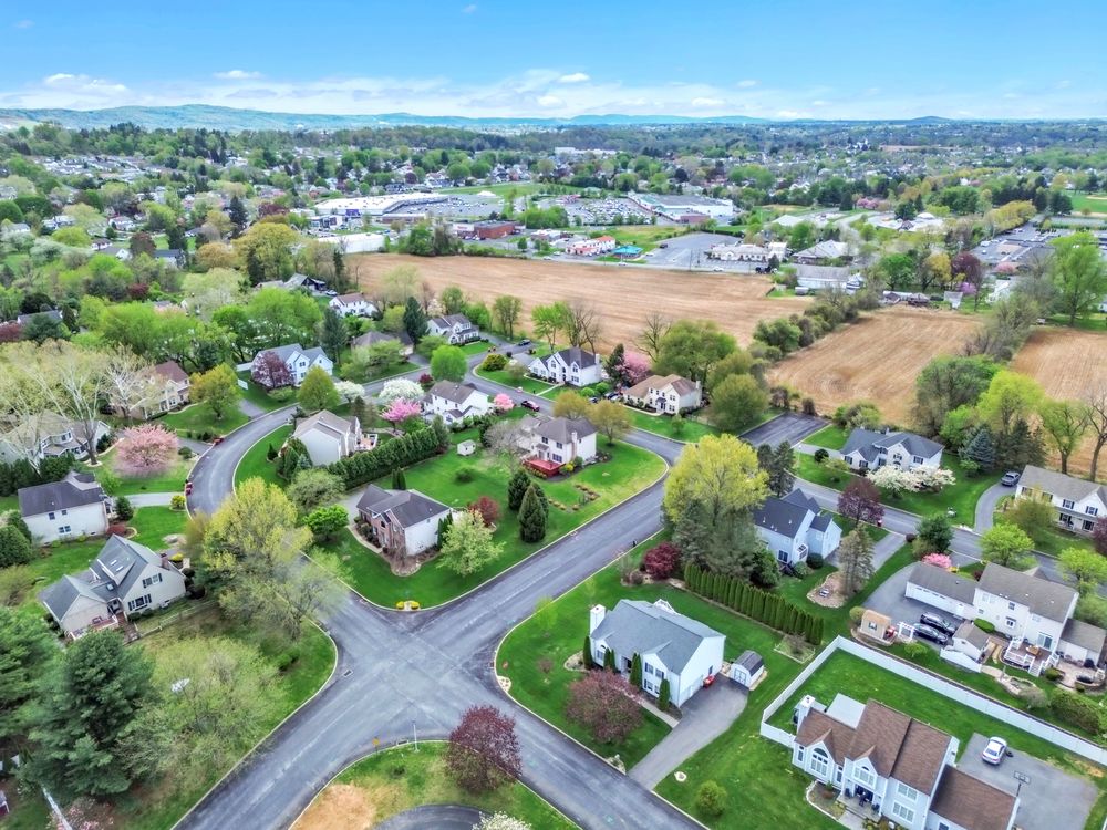 Aerial drone view of a peaceful suburban neighborhood with lush green trees, residential homes, and open fields under a bright blue sky.