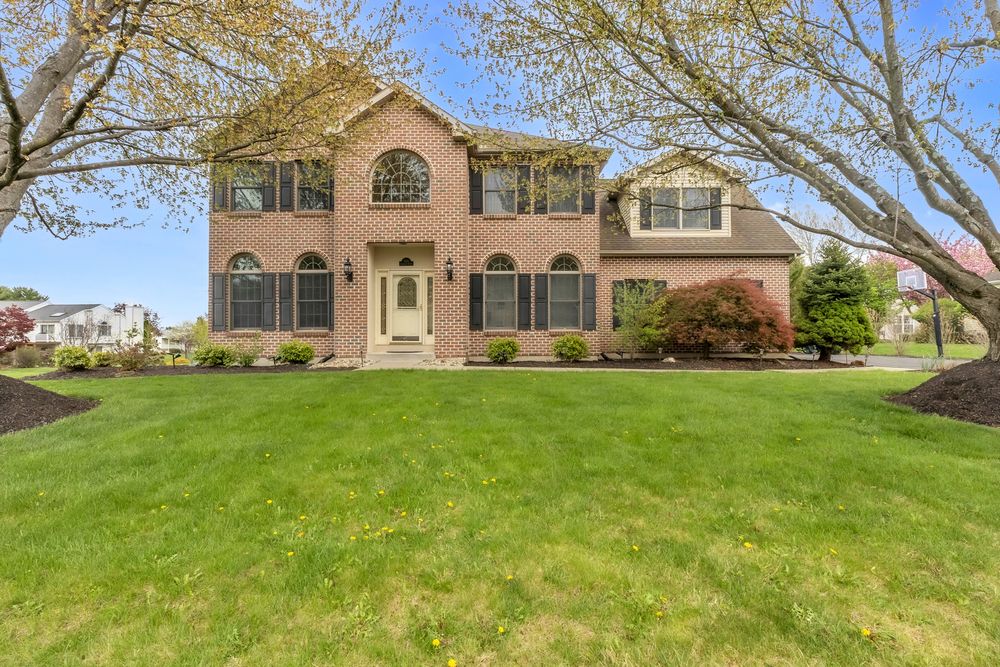 Two-story brick colonial home with arched windows, lush green lawn, and mature trees in a suburban neighborhood.