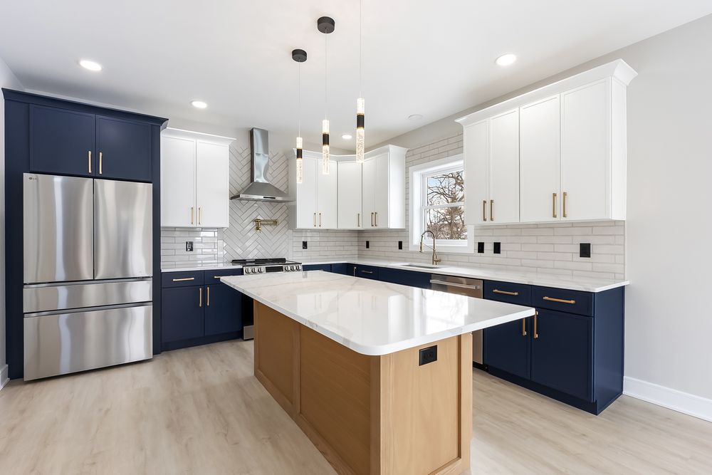 Modern kitchen with navy blue and white cabinets, stainless steel fridge, quartz island, and pendant lights.