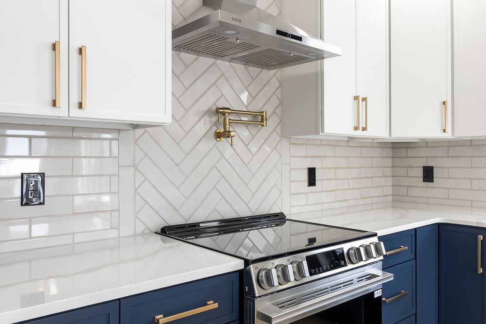 Modern white kitchen with navy blue lower cabinets, white herringbone backsplash, and gold hardware accents.