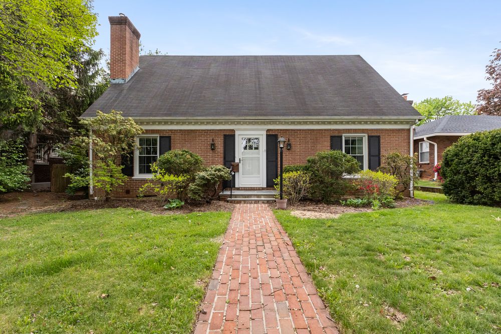Charming brick Cape Cod home with manicured lawn, black shutters, white front door, and brick pathway leading to entrance.