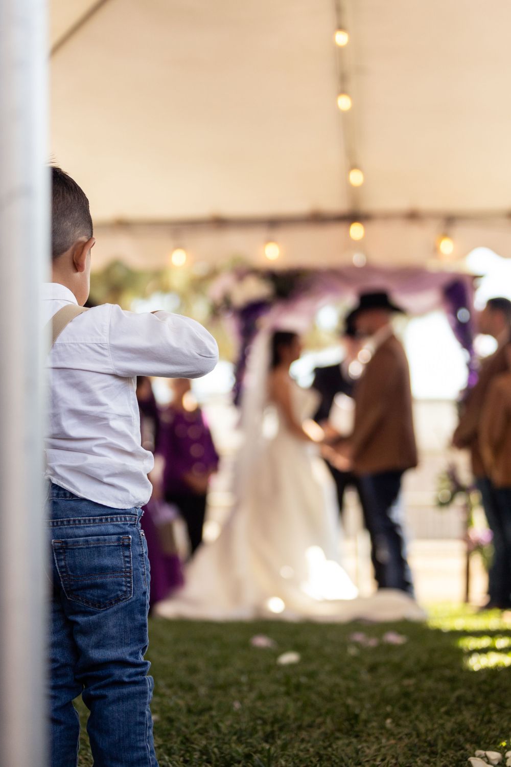 A young child watches a wedding ceremony from the sidelines under a tent with string lights.