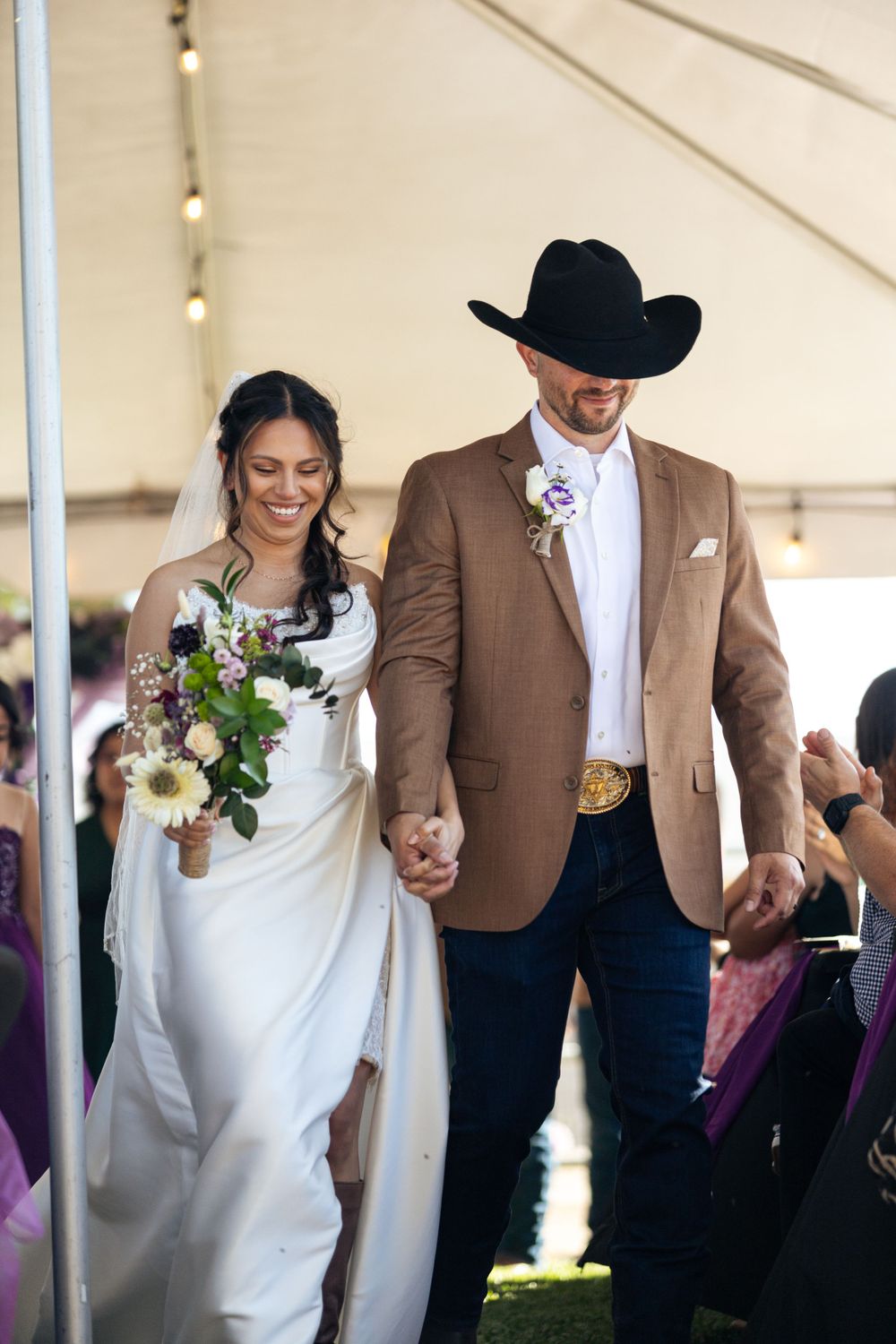 A couple walks down the aisle after their wedding ceremony, with the groom wearing a cowboy hat.