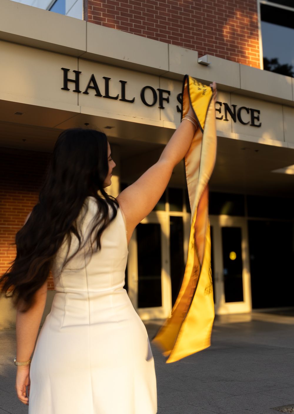 A student in white clothing holds up a yellow graduation stole against a brick building exterior.