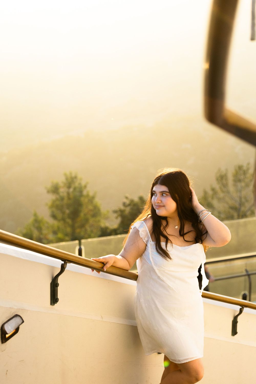 Young woman in white dress leaning on golden railing at sunset with scenic mountain view.