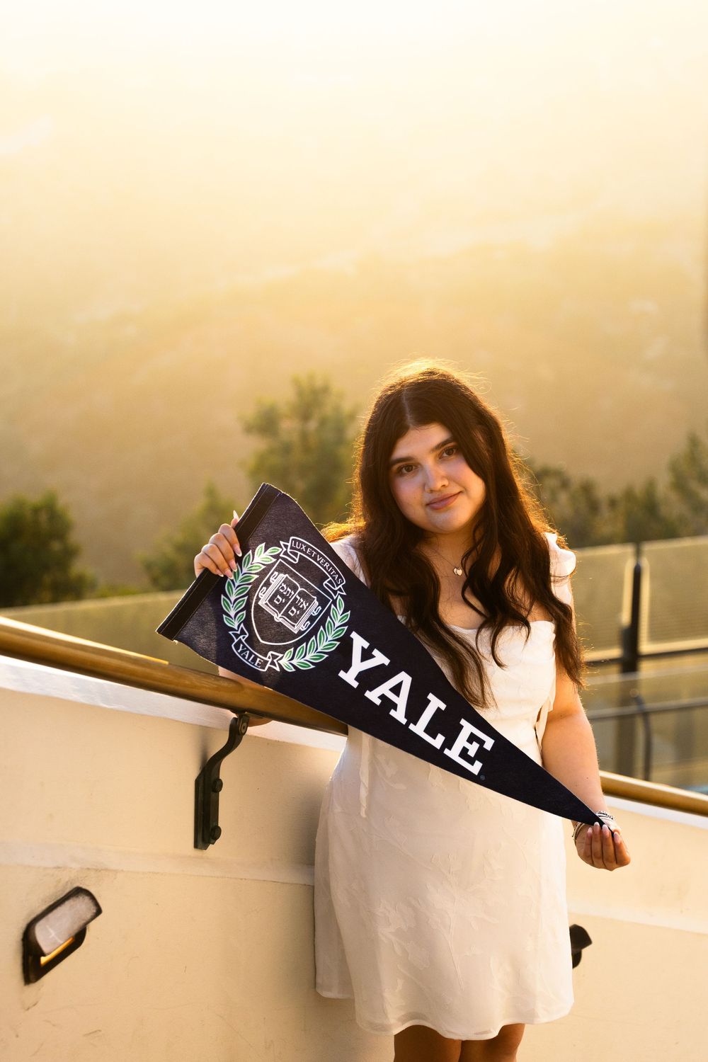 Young woman in white dress holding a Yale University pennant flag outdoors at golden hour.