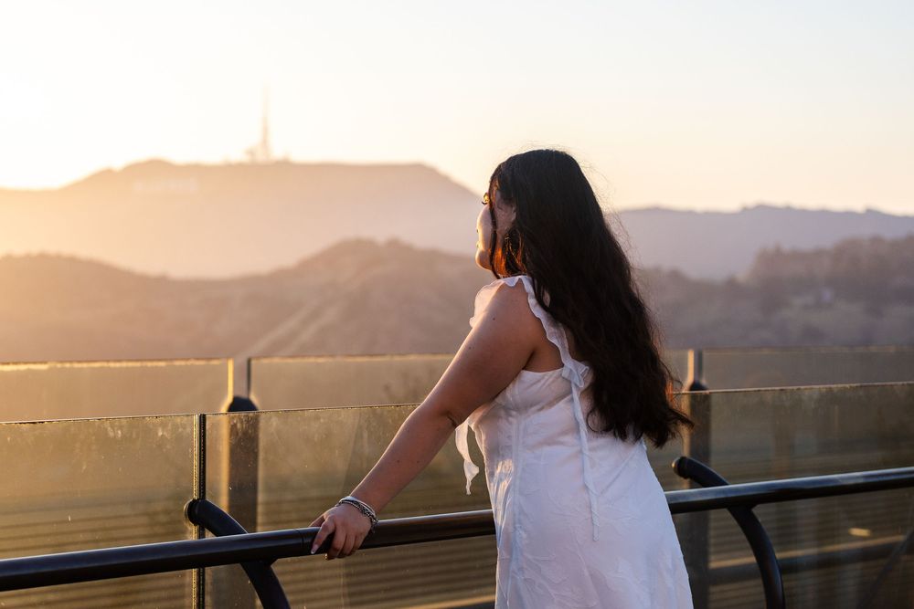 Young woman in white dress gazes at sunset over Hollywood Hills from a scenic overlook balcony.
