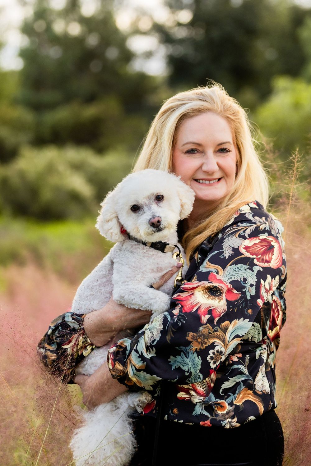 A white Bichon Frise puppy is held by someone wearing a floral print top in an outdoor nature setting.