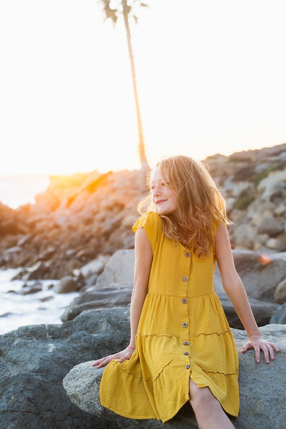 A person in a flowing yellow sundress sits gracefully on coastal rocks during golden hour sunset.