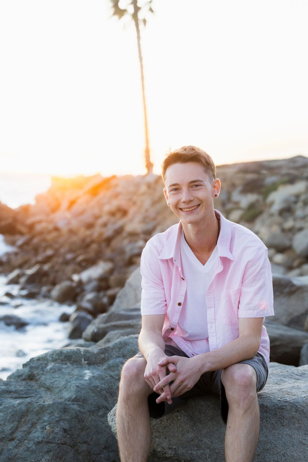 A person in a pink shirt sits on coastal rocks at sunset while smiling at the camera.