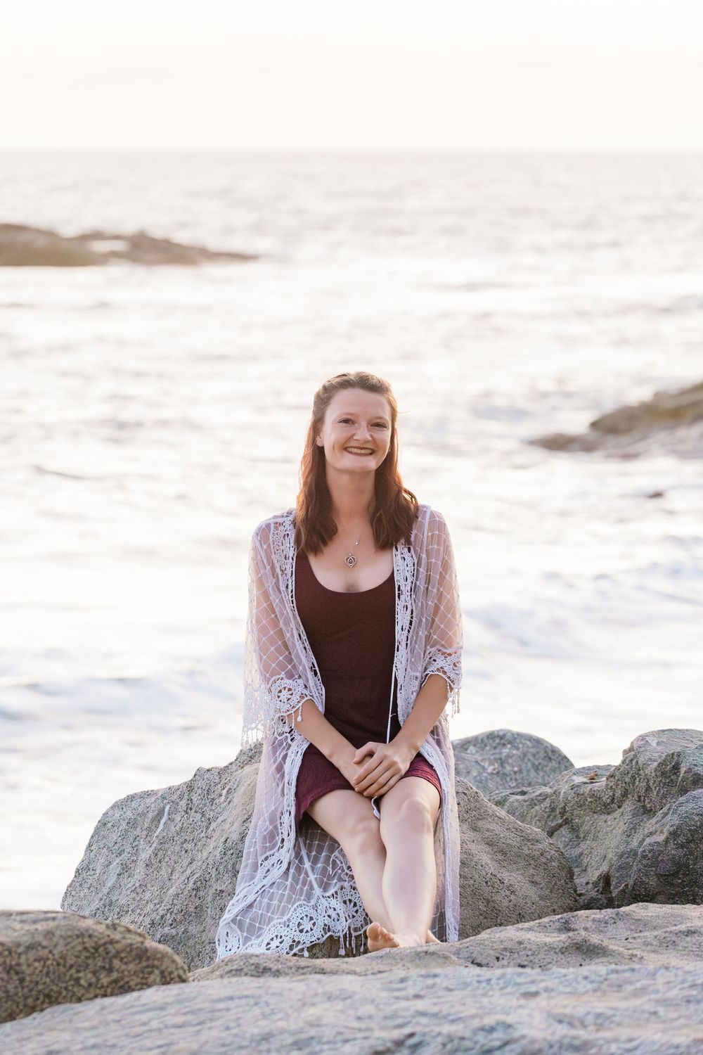 A person in a brown dress and white lace cardigan sits on coastal rocks at sunset near the ocean waves.
