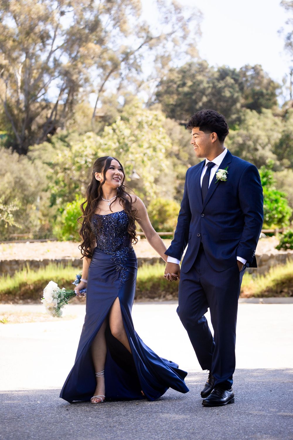 A couple in matching navy blue prom attire hold hands and smile at each other outdoors.