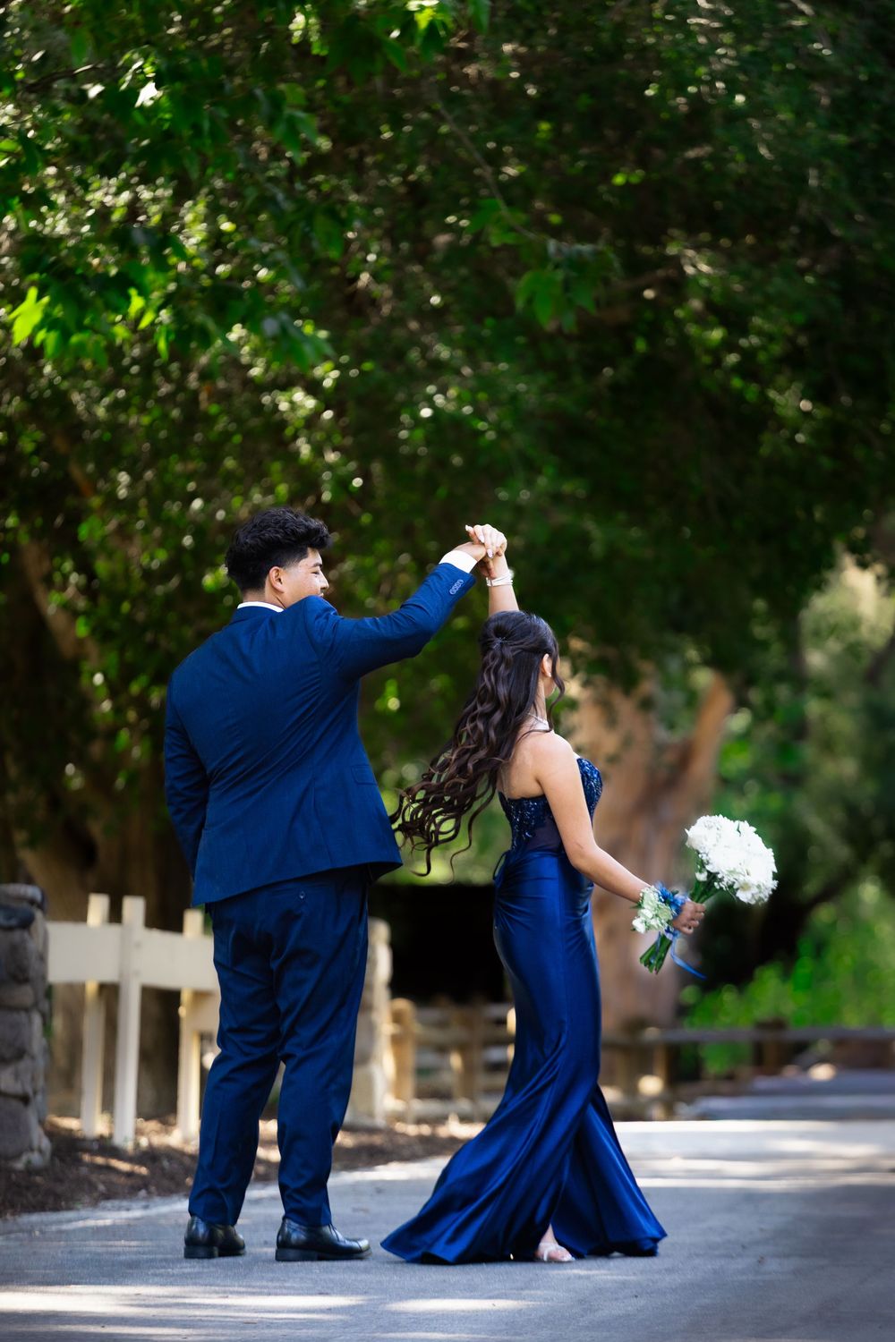 Couple in matching navy blue formal wear dancing outdoors, woman holding white bouquet, surrounded by lush green trees.