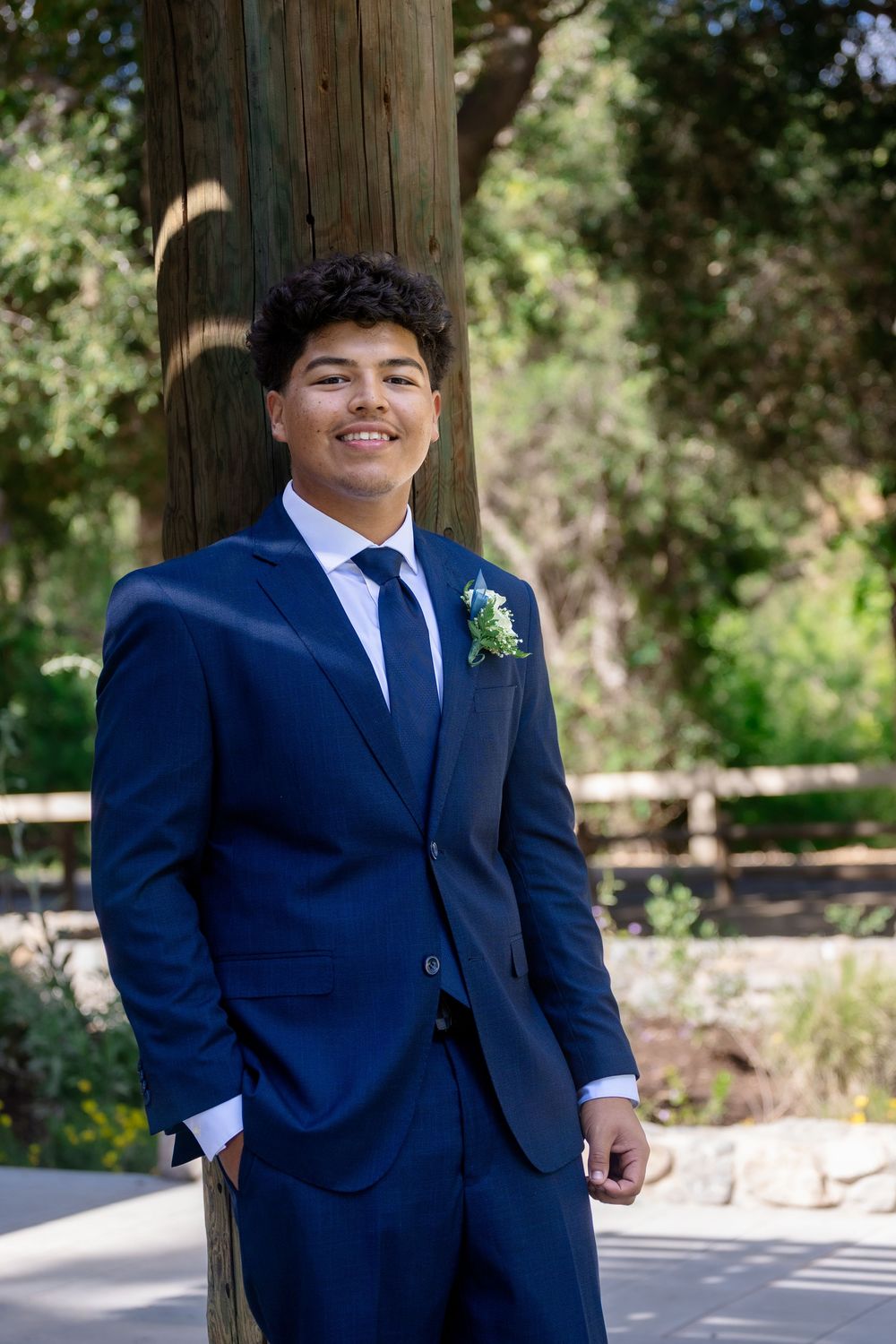 Young man in navy blue suit with boutonniere smiling outdoors near tree at formal event.