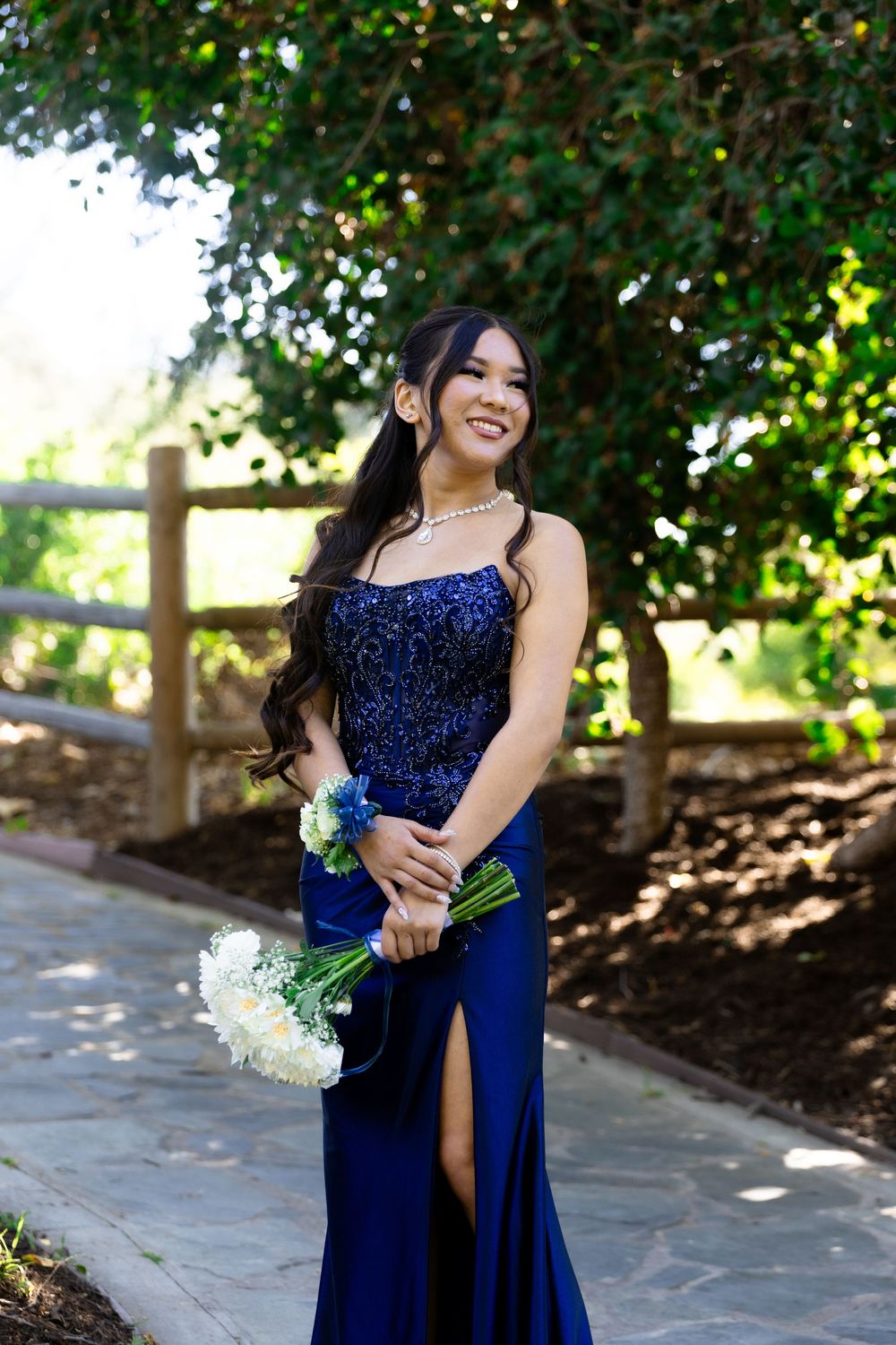 Young woman in navy blue sequin prom gown holding white and blue bouquet, posing outdoors near wooden fence.