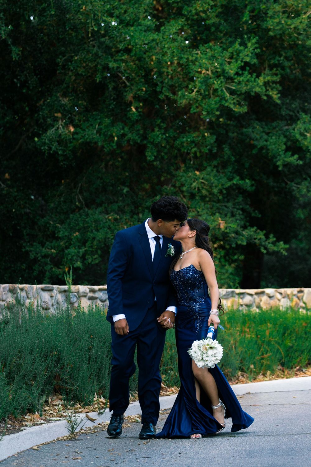 Couple in matching navy blue formal wear sharing a kiss outdoors, woman holding white bouquet, lush green trees behind.