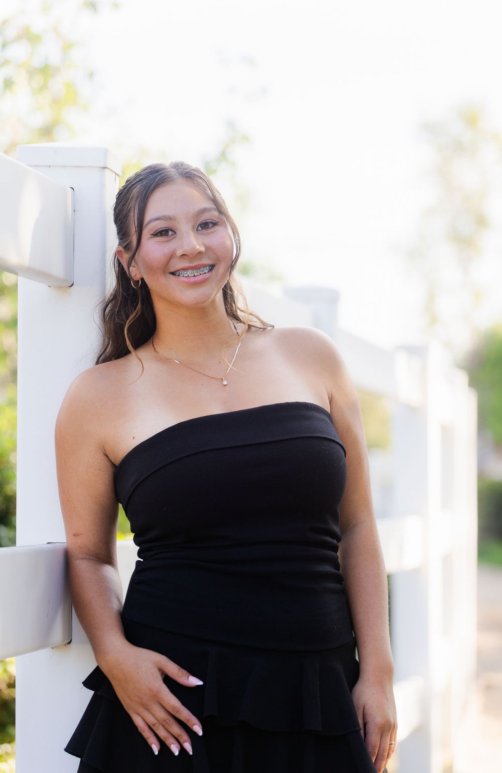 A person in a strapless black dress poses confidently outdoors against a light-colored wall.