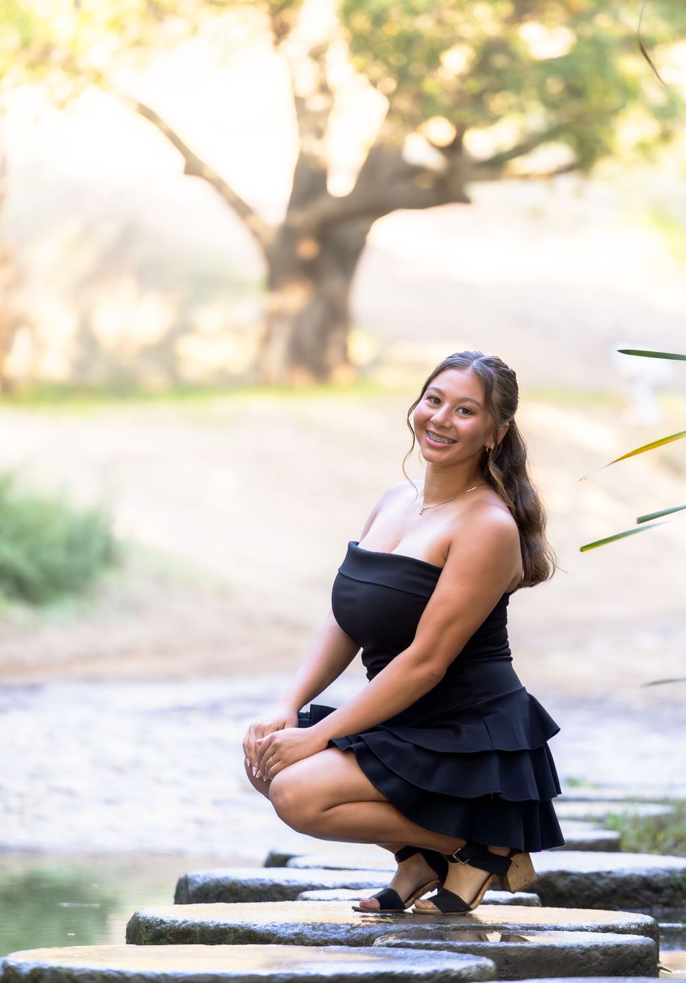 Woman in black dress poses gracefully on rocks near a serene pond in a natural outdoor setting.