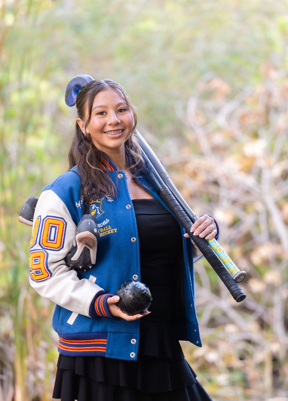High school student in blue and orange varsity jacket holds softball gear in outdoor portrait setting.