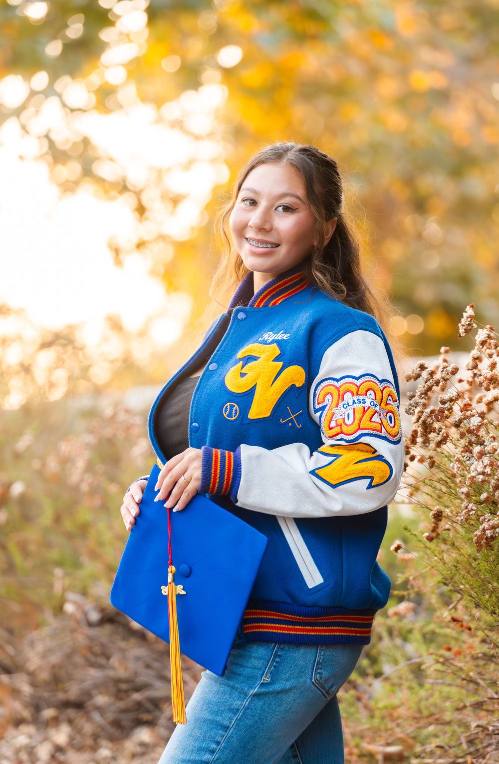 A person wearing a blue and orange varsity jacket poses outdoors against a blurred autumn background.