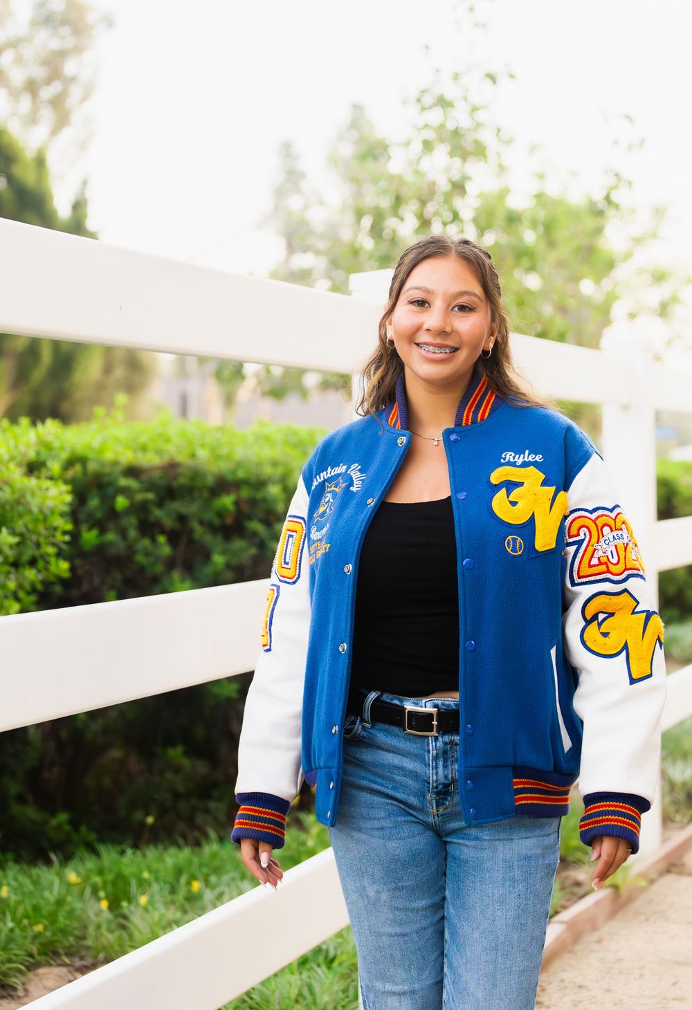 A person wearing a blue varsity jacket and jeans poses against a white fence with greenery in the background.