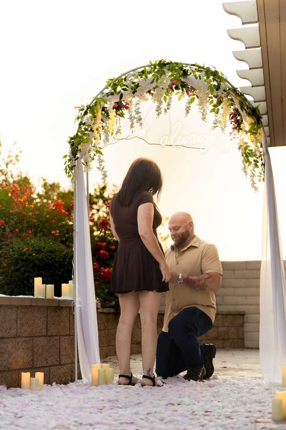 Man kneeling on one knee proposing to woman under floral arch with candles and rose petals at sunset.