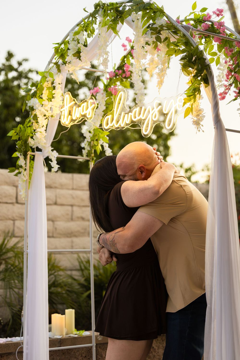 Couple embracing under a floral arch with neon 'always' sign and candles during an outdoor proposal.