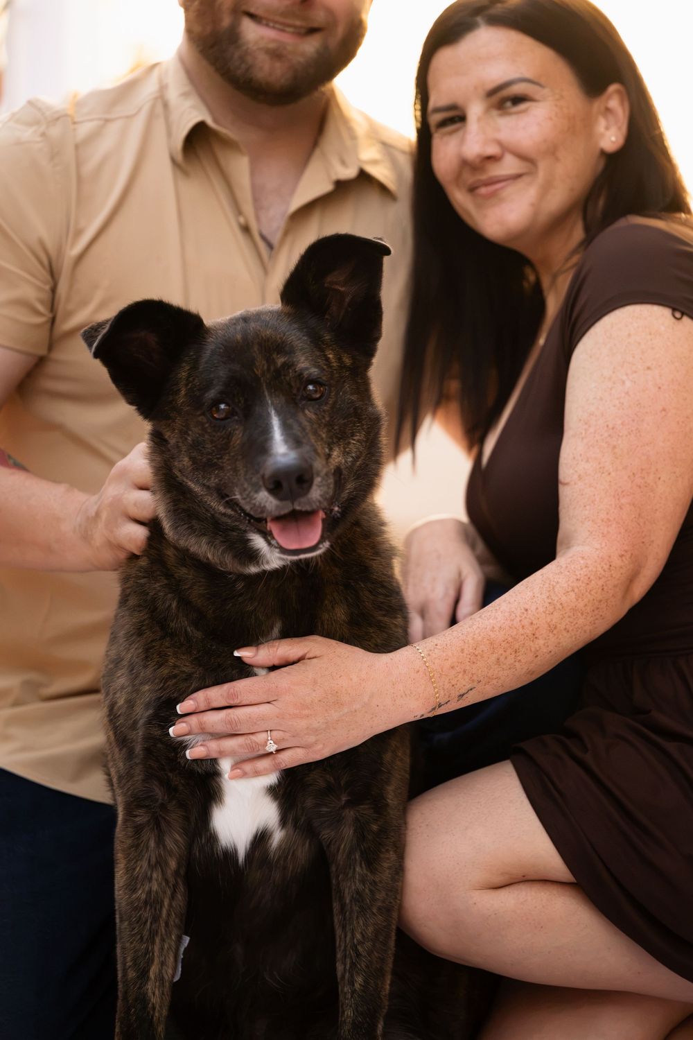 A happy couple smiling and posing with their brindle dog, gently petting it in a warm indoor setting.