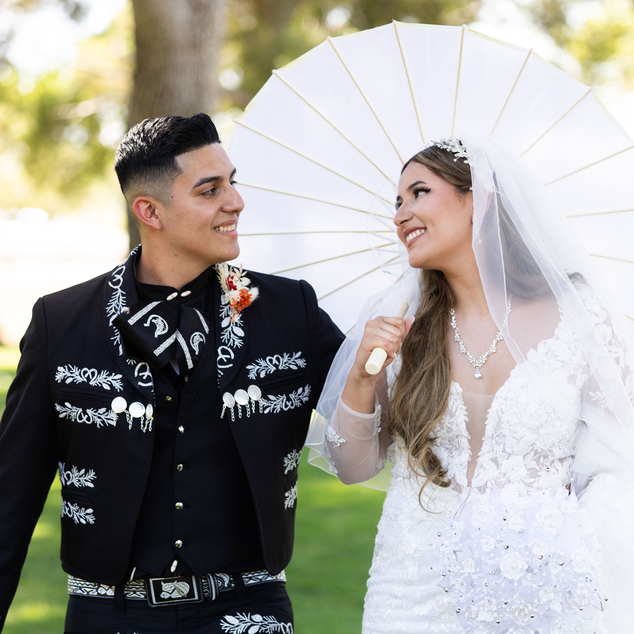 Bride in white gown holding parasol and groom in black charro suit smiling at each other outdoors.