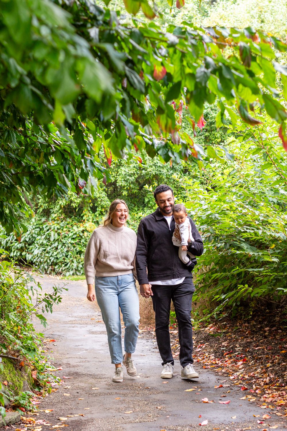 Couple walking together on leaf-covered pathway surrounded by lush greenery.