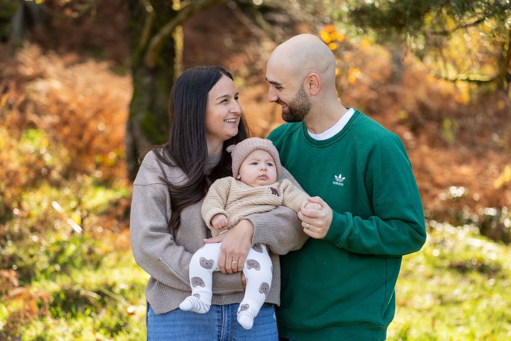 A couple holds their baby outdoors during autumn with warm golden foliage in the background.